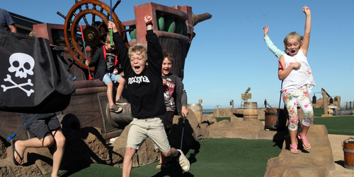 Children jumping up and down in happiness next to a pirate ship.  Theyre enjoying adventure gold at Pirate Golf and Splash Pad.