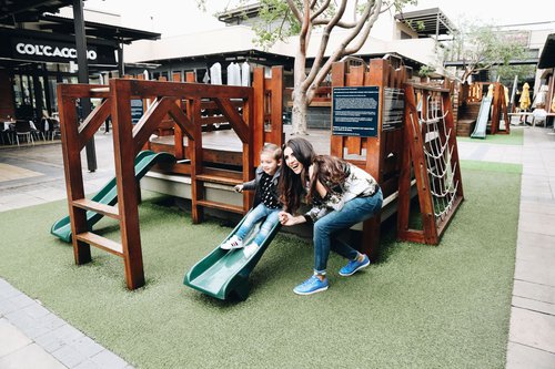 A mom and her daughter are posing for a photo as the child goes down a slide off of a jungle gym in the middle of the Willowbridge Shopping Centre