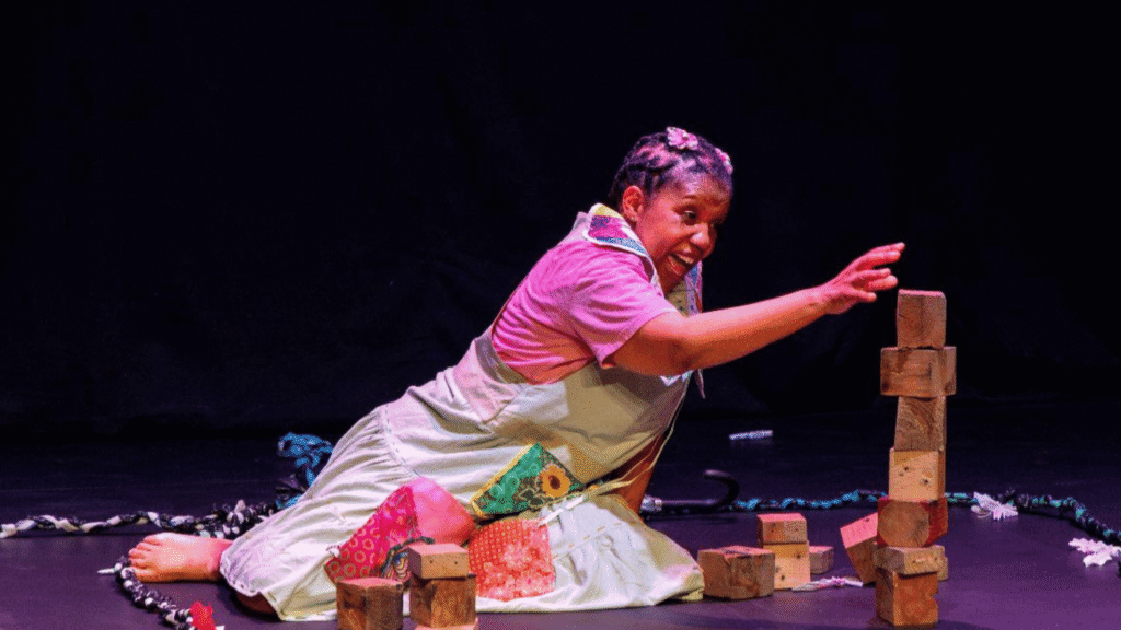 A performer in Serurubele reaches toward a tower of wooden blocks during a colourful stage performance at the Cradle of Creativity Festival 2026.