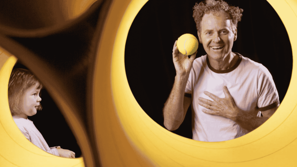 A performer in The Boy and the Ball holds up a yellow ball while interacting with a young child during a playful theatre performance at the Cradle of Creativity Festival 2026.